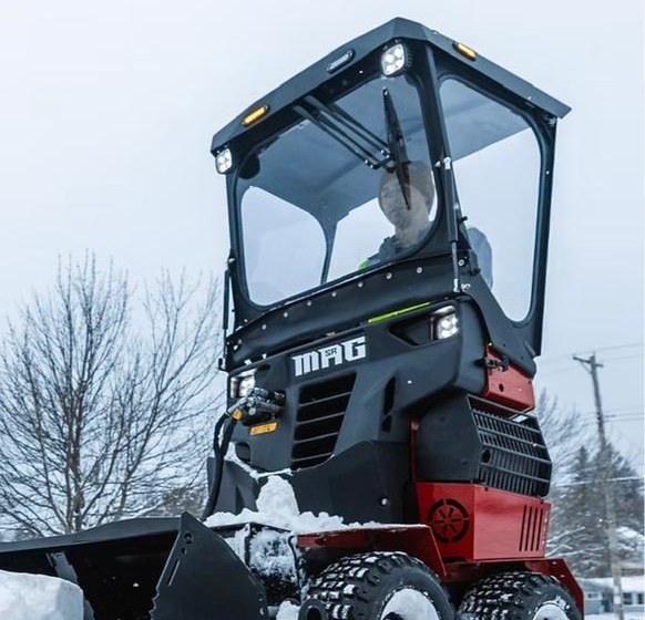 Articulated sidewalk machine on chained tyres, fitted with a plow blade, clearing a walkway